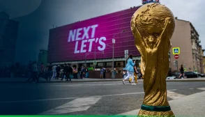 Uma foto do troféu da copa do mundo na calçada da rua, com um letreiro digital gigante ao fundo do outro lado da rua escrito: "Next Let's"