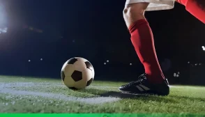 Jogador de futebol prestes a chutar uma bola em campo iluminado durante a noite; ele veste meião vermelho, chuteira preta e uniforme branco, com foco nos pés e na bola posicionada sobre a grama.
