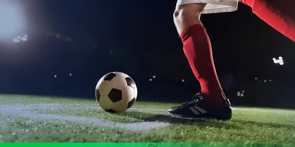Jogador de futebol prestes a chutar uma bola em campo iluminado durante a noite; ele veste meião vermelho, chuteira preta e uniforme branco, com foco nos pés e na bola posicionada sobre a grama.