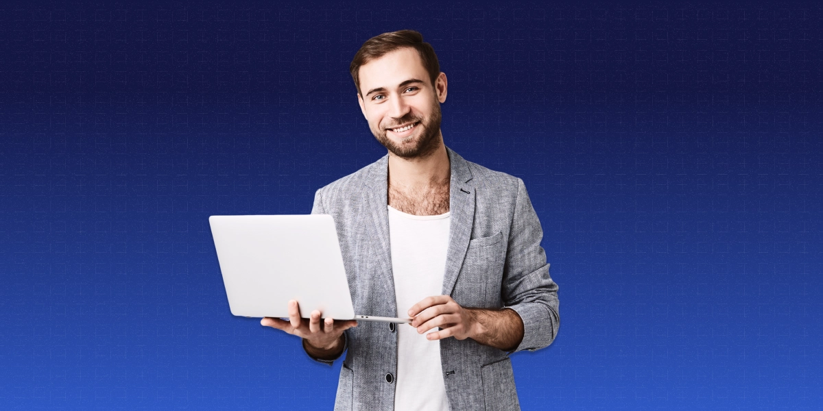 imagem mostra homem sorrindo ao segurar um notebook. Ele veste camiseta branca e um blazer cinza.