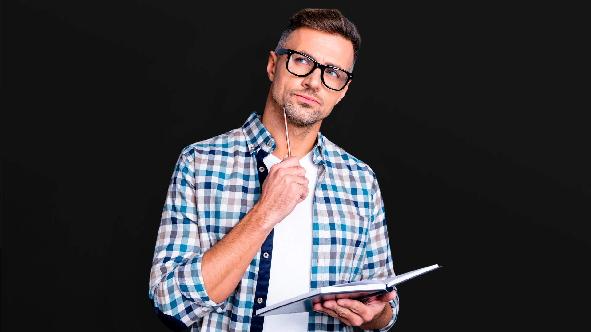 Um homem adulto, de pele clara, usando óculos de aro preto e uma camisa xadrez azul e branca sobre uma camiseta branca. Ele está segurando um pequeno caderno preto e um lápis na mão direita, com o lápis apoiado no queixo. Sua expressão é pensativa ou atenta, e o fundo é preto.