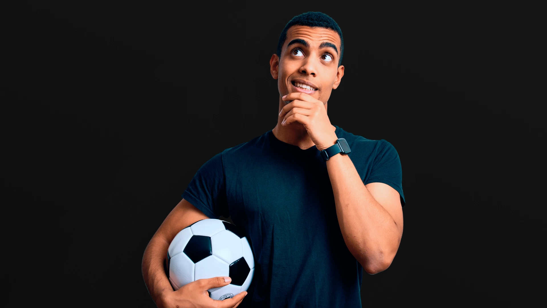 Um homem jovem de pele escura, com camiseta preta e segurando uma bola de futebol no braço esquerdo. Ele tem uma expressão pensativa, com a mão direita no queixo e olhando para cima, para a direita. A imagem está em um fundo escuro.