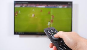 Imagem mostra um jogo de futebol e uma mão segundo um controle remoto