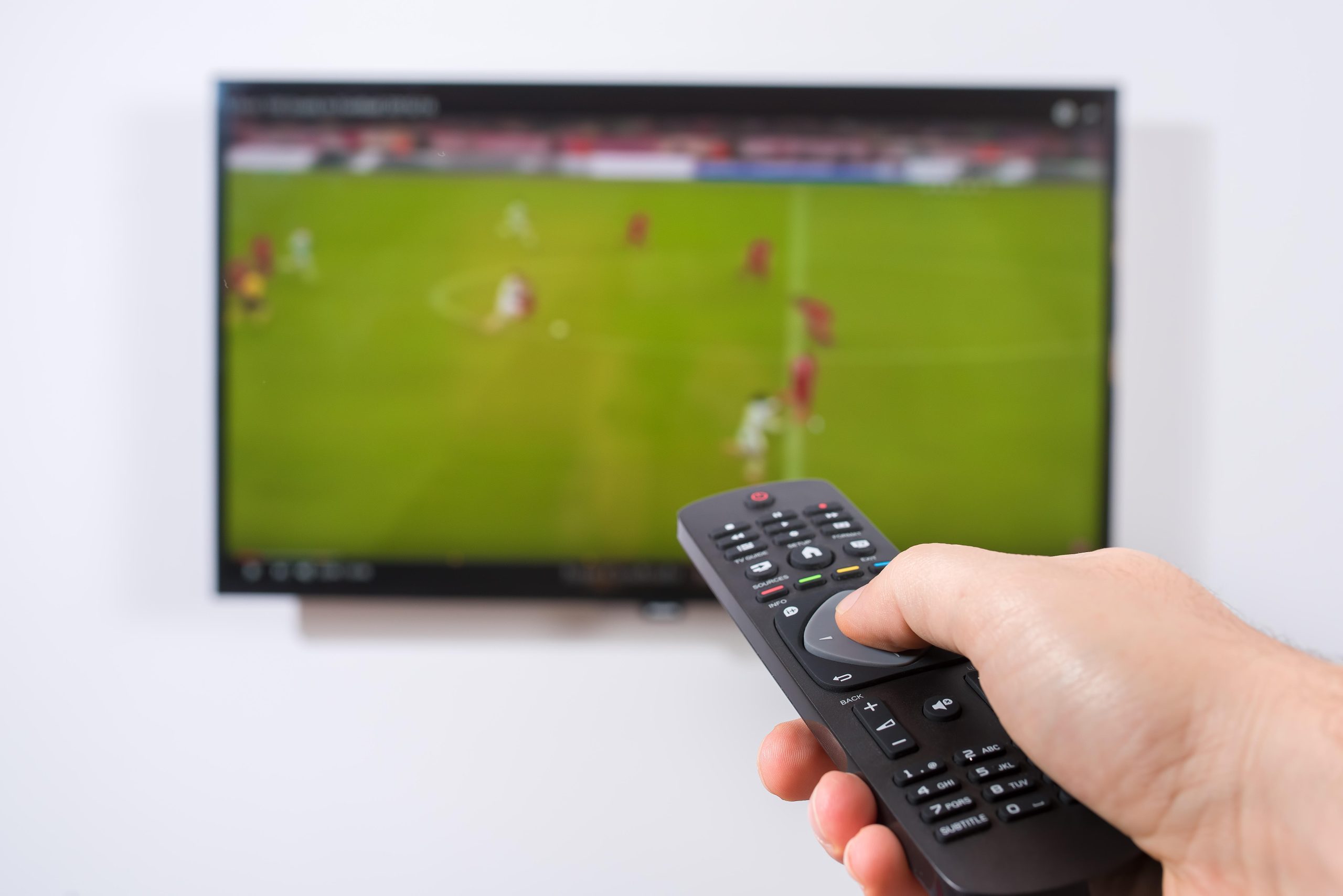 Imagem mostra um jogo de futebol e uma mão segundo um controle remoto