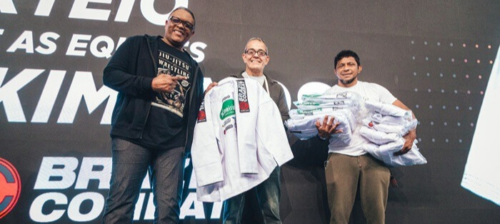 Três homens da Brazil Combat no palco de um evento da CBJJD, segurando e entregando quimonos brancos de Jiu-Jitsu com diversos logotipos de patrocinadores