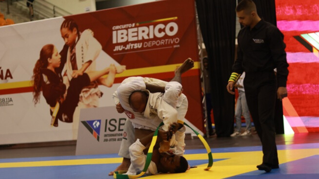 Luta de Jiu-Jitsu Brasileiro (BJJ) no Circuito Ibérico ISBJJA, mostrando um competidor por cima tentando uma finalização contra o adversário no tatame sob supervisão do árbitro. A cidade de Coimbra