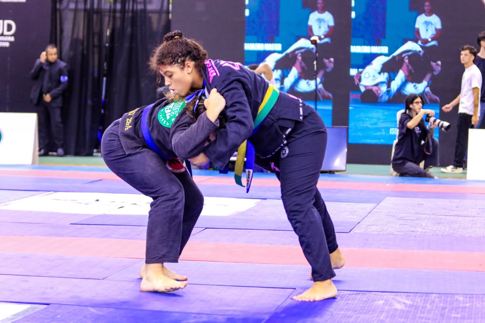 Duas atletas de Jiu-Jitsu em combate em pé durante Circuito Mineirinho de Jiu-Jitsu, segurando os quimonos uma da outra em busca de uma queda no tatame azul.